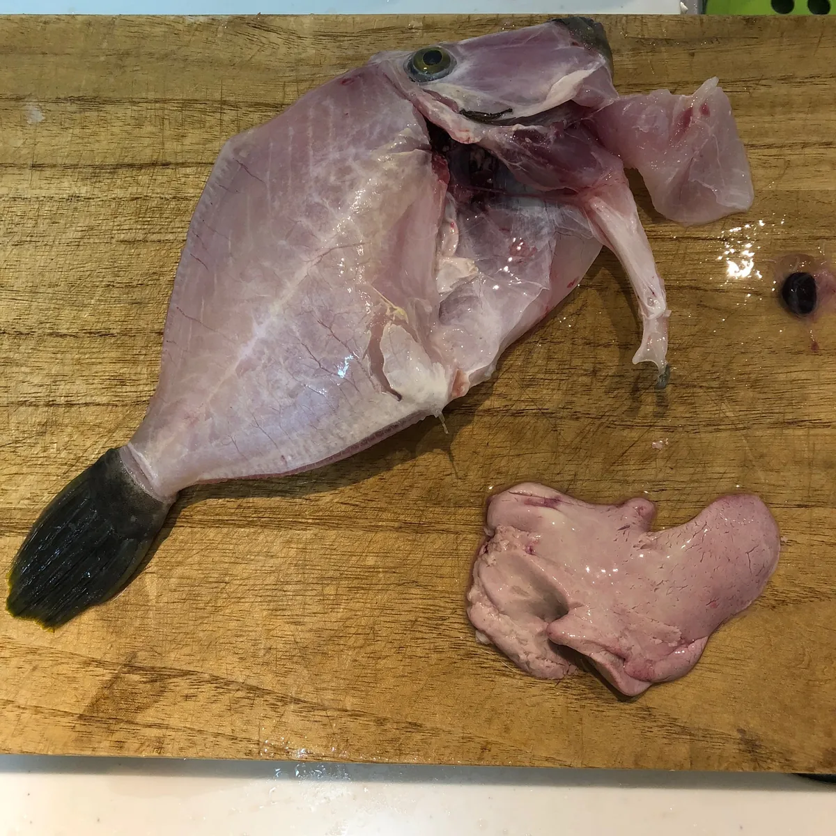 木製のまな板の左側に、皮を剥かれ白っぽいピンク色をした魚体(目と尾が確認できる)、右側に血管の筋が透ける淡いピンク色で不定形の肝臓が置かれている。上部には胆嚢とみられる黒い粒が付着